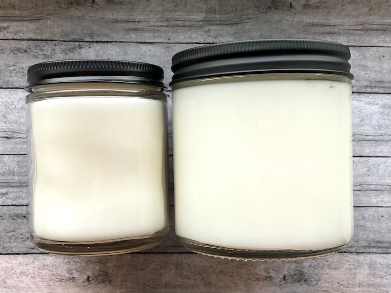 May include: Two glass candle jars with black lids. The jars are filled with white wax and are sitting on a wooden surface.