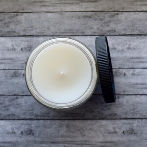 May include: A white candle in a clear glass jar with a black lid. The candle is unlit and has a single wick.