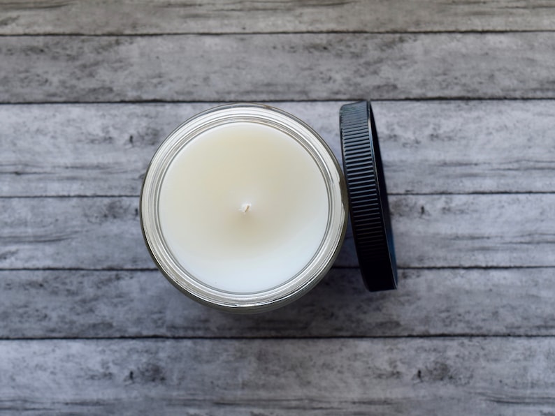 May include: A white candle in a clear glass jar with a black lid. The candle is unlit and has a single wick.