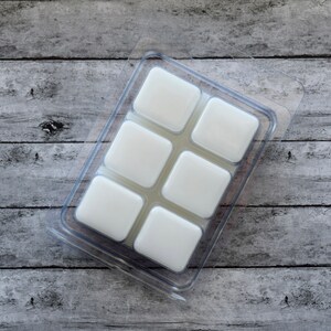 May include: A clear plastic mold with six square compartments filled with white wax melts. The mold is sitting on a wooden surface.