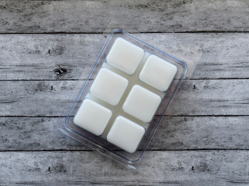 May include: A clear plastic mold with six square compartments filled with white wax melts. The mold is sitting on a wooden surface.