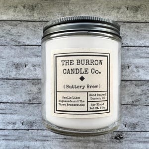 May include: A clear glass candle jar with a black metal lid containing white wax. The label reads: THE BURROW CANDLE Co. { Buttery Brew } Smells Like: Hogsmeade and The Three Broomsticks. Hand Poured Duncan, OK. Soy Blend Net Wt. 8 Oz.