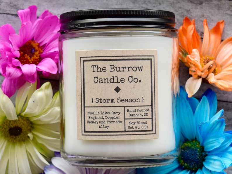 May include: A glass jar candle with a white label that reads "The Burrow Candle Co. {Storm Season}". The label also includes the text "Smells Like: Gary England, Doppler Radar, and Tornado Alley", "Hand Poured Duncan, OK", "Soy Blend", and "Net Wt. 8 Oz".