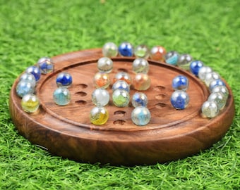Puede incluir: Un juego de mesa de solitario de madera redondo con una serie de hendiduras circulares dispuestas en forma de cruz. El tablero está poblado con varias canicas de vidrio de colores, incluyendo tonos azules, amarillos, transparentes y naranjas.