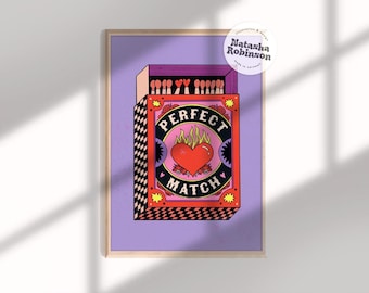 May include: Framed art print with a matchbox design. The artwork features the words "Perfect Match" within a heart-shaped emblem. The matchbox is red, black, and pink, set against a purple background, and framed in wood.