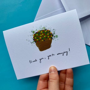 May include: A white card with a hand-painted illustration of a potted plant with colorful flowers. The text "thank you, you're amazing!" is written in black ink.