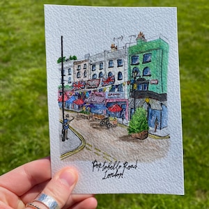 Könnte beinhalten: Aquarellmalerei der Portobello Road in London. Das Kunstwerk zeigt eine Straßenszene mit Marktständen, Union-Jack-Flaggen und einem Musiker. Die Gebäude sind in Grau- und Grüntönen gehalten, mit bunten Markisen.