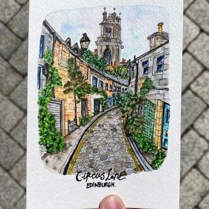 May include: Watercolor painting of a narrow cobblestone street in Edinburgh, Scotland. The street is lined with old buildings and a tall church tower is visible in the background. The painting is signed "Circus Lane, Edinburgh."