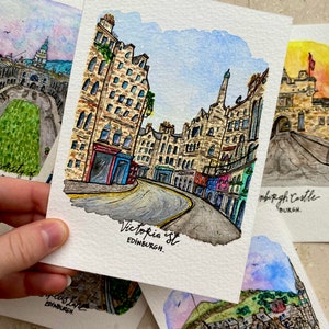 May include: Watercolor painting of a street scene in Edinburgh, Scotland. The street is lined with tall, narrow buildings with stone facades. The street is curved and there is a blue sky with white clouds in the background. The text "Victoria St. EDINBURGH." is written at the bottom of the painting.