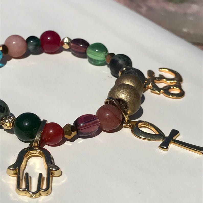 May include: A beaded bracelet featuring a variety of colorful beads in shades of pink, green, red, and gold. The bracelet includes gold charms of a hand, an ankh, and the word "love". The beads are strung together on an elastic band.