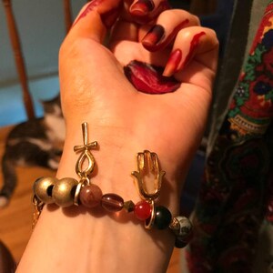 May include: Gold-toned beaded bracelet with an ankh and Hamsa hand charm. The bracelet features a mix of gold, brown, red, and green beads. The ankh charm is a symbol of life, and the Hamsa hand is a symbol of protection.