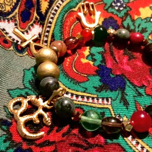 May include: A beaded bracelet with gold-colored charms and a variety of colorful beads. The charms include a cross, a hand, and an Om symbol. The beads are in shades of green, red, brown, and gold.