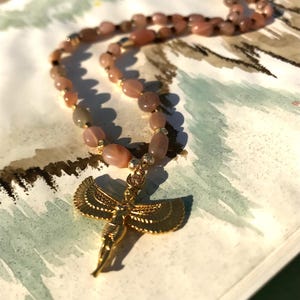 以下が含まれることがあります： ピーチ色の宝石ビーズと金色のペンダントが付いたネックレス。ペンダントは翼のある姿をしています。ネックレスは模様のある表面に表示されています。