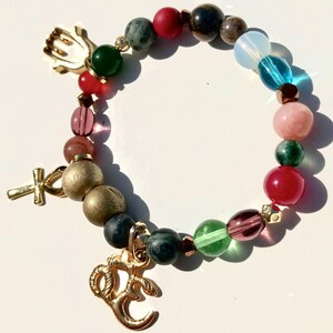 May include: A beaded bracelet featuring a variety of colorful beads in shades of red, green, pink, and blue. The bracelet includes gold-colored charms, including a hand, a cross, and an Om symbol. The beads are strung together on an elastic band.
