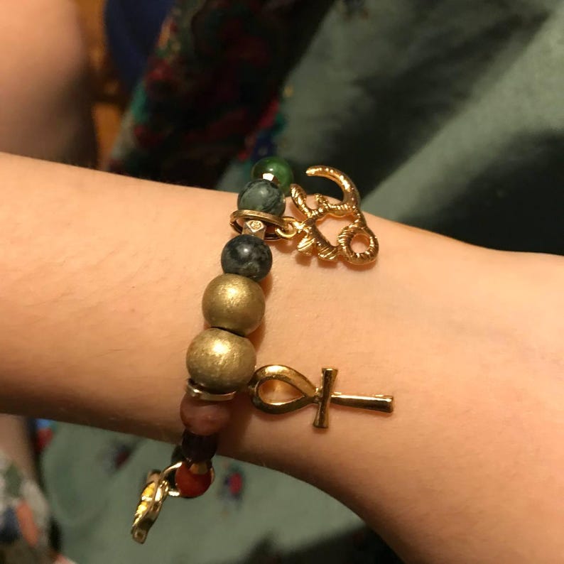 May include: A beaded bracelet with gold-colored charms. The bracelet features a mix of colorful beads, including green, black, gold, and red. Two gold charms are attached: an Ankh symbol and a decorative design.