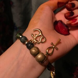 May include: A beaded bracelet with gold-colored charms, including an Om symbol and an Ankh cross. The bracelet features a mix of dark green, black, and gold-colored beads. The background is a hand with henna designs.