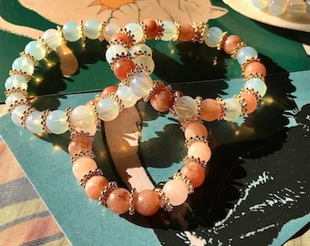 May include: Two beaded bracelets, one with light pink and white beads and the other with white and light blue beads. Both bracelets have gold accents.