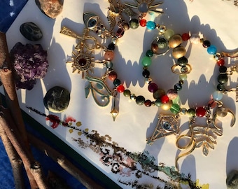 May include: A collection of beaded bracelets and charms with gold-colored accents. The bracelets feature a variety of colorful beads, including red, green, and gold. Decorative charms include sunbursts, ankhs, and eye symbols. The image also includes small stones and twigs.