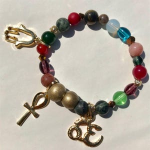 May include: A beaded bracelet with a variety of colorful beads, including red, green, blue, and gold. The bracelet features gold charms of a hand, an ankh, and the Om symbol. The beads are strung on an elastic cord.