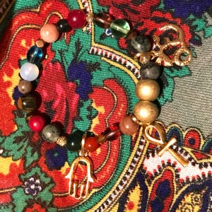 May include: A beaded bracelet featuring a variety of colorful gemstones and gold-toned charms. The bracelet includes a gold-toned ankh and a Hamsa hand charm. The beads are in shades of red, green, brown, and gold.
