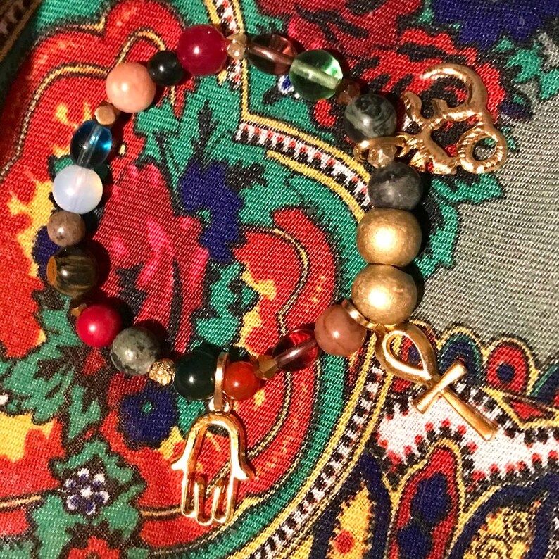 May include: A beaded bracelet featuring a variety of colorful gemstones and gold-toned charms. The bracelet includes a gold-toned ankh and a Hamsa hand charm. The beads are in shades of red, green, brown, and gold.