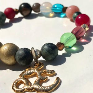 May include: A beaded bracelet featuring a variety of colorful beads in shades of gold, green, blue, pink, and red. The bracelet includes a gold-colored charm with an ornate design. The beads are of varying sizes and textures.