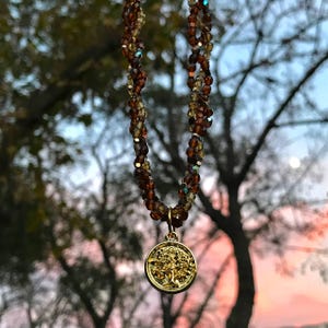 May include: A necklace with brown and amber-coloured beads and a gold-coloured pendant. The pendant has a tree design. The necklace is suspended against a blurred backdrop of trees and a sunset. The beads are measured in centimetres.