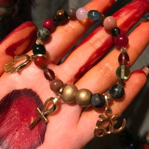 May include: A beaded bracelet with a variety of colorful beads in shades of red, green, blue, and brown. The bracelet features gold-colored charms, including a hand, an ankh, and an Om symbol. The bracelet is displayed on a hand.