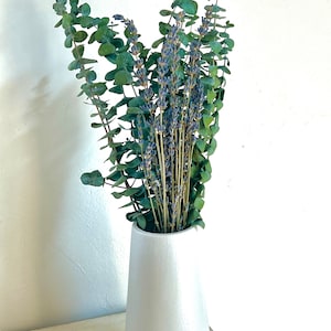 A bundle of bright green eucalyptus and bright purple lavender in a white cylindrical vase sitting on a white table as home decor.