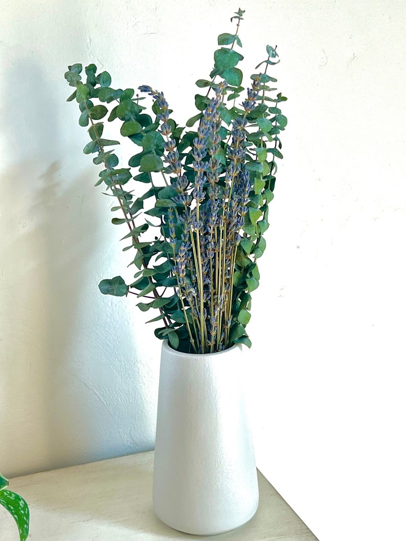 A bundle of bright green eucalyptus and bright purple lavender in a white cylindrical vase sitting on a white table as home decor.