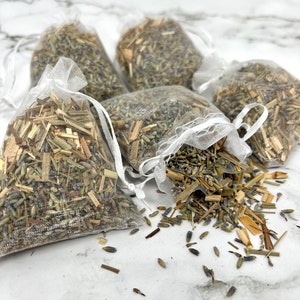 May include: Five small white mesh bags filled with dried lavender and lemongrass. The bags are tied with white ribbon and are scattered on a white marble surface.