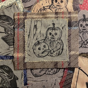 May include: A patchwork of fabric squares featuring black and white pumpkin-themed illustrations. The squares are sewn together with various fabrics, including plaid and floral patterns. The pumpkin designs have a folk art style.
