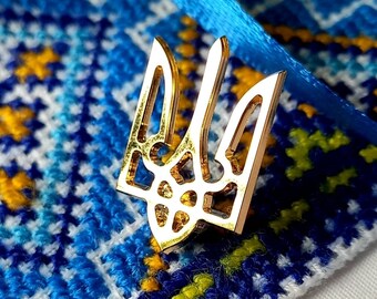 May include: A gold-colored metal trident, the national symbol of Ukraine, is placed on top of a blue and yellow embroidered fabric. The fabric has a traditional Ukrainian pattern.
