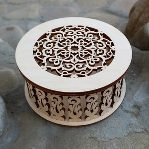 May include: A round wooden box with a decorative, intricate, laser-cut design. The box has a lid with a circular cutout revealing the design inside. The box is made of light-colored wood.