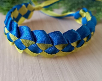 May include: Braided bracelet featuring a blue and yellow woven design. The bracelet is made of satin ribbon and has a woven pattern. The ends of the ribbon extend from the bracelet. The bracelet is on a light wood surface.