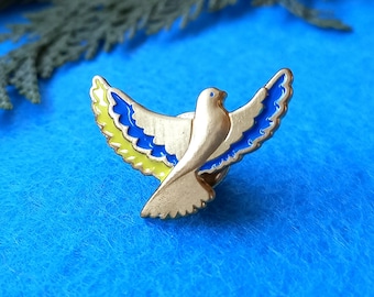 May include: A gold-colored metal peace dove pin with blue and yellow enamel wings. The dove is a symbol of peace and hope.