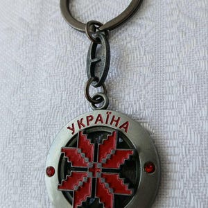 May include: A circular metal keychain featuring a red, eight-pointed geometric star emblem at the center. The outer rim is engraved with the text "УКРАЇНА" and "UKRAINE" in red, accented by two small red circular stones on the left and right sides.