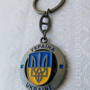 May include: A circular metal keychain featuring a blue and yellow shield with the Ukrainian trident emblem. The outer ring displays the text "УКРАЇНА" and "UKRAINE" with small blue star accents on each side. The keychain includes a metal ring.