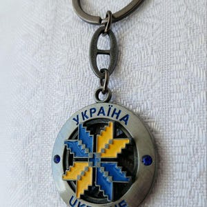 May include: A circular metal keychain featuring a gunmetal finish and a central eight-pointed star design in blue and yellow enamel. The outer ring displays the text "УКРАЇНА" and "UKRAINE" in blue, accented by two small blue rhinestones on the sides.