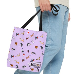 K&ouml;nnte beinhalten: Eine lavendelfarbene Tragetasche mit einem sich wiederholenden Muster von verspielten Hundeillustrationen in verschiedenen Posen. Die Tasche hat schwarze Griffe und den Text "WET LAB CREATIVE" in einem wei&szlig;en Feld unten rechts.