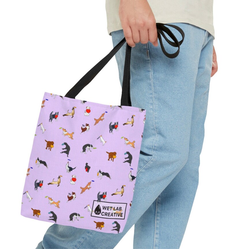 K&ouml;nnte beinhalten: Eine lavendelfarbene Tragetasche mit einem sich wiederholenden Muster von verspielten Hundeillustrationen in verschiedenen Posen. Die Tasche hat schwarze Griffe und den Text "WET LAB CREATIVE" in einem wei&szlig;en Feld unten rechts.