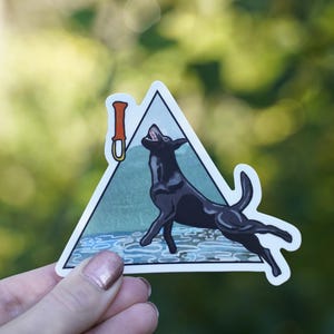Op de afbeelding: Een sticker met een zwarte hond die omhoog springt in een wit omrandde driehoek. De driehoek bevat een berglandschap en een klimgereedschap. De hond is het middelpunt, met zijn silhouet en het klimgereedschap in oranje.