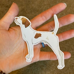 May include: A sticker featuring a white dog with brown markings. The dog is in a side profile, with its head tilted upwards. The sticker has a white border and is held in a person's hand.