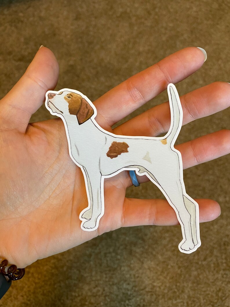 May include: A sticker featuring a white dog with brown markings. The dog is in a side profile, with its head tilted upwards. The sticker has a white border and is held in a person's hand.