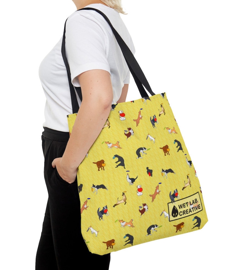 K&ouml;nnte beinhalten: Gelbe Tragetasche mit einem sich wiederholenden Muster von verspielten Hunden in verschiedenen Posen. Die Tasche hat schwarze Tr&auml;ger und den Text "WET LAB CREATIVE". Die Tasche wird &uuml;ber der Schulter getragen.