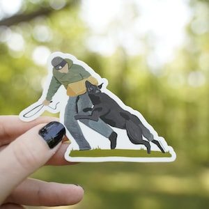 May include: A sticker featuring a dog training scene. The illustration depicts a person in a green jacket and gray pants with a black dog. The dog is jumping towards the person. The sticker is held by a hand with black nail polish.