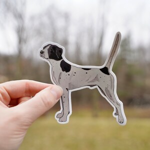 May include: A sticker featuring a stylized illustration of a dog, primarily white with black markings on its head and back. The dog is depicted in a side profile with its tail raised. The sticker is held against a blurred outdoor background.