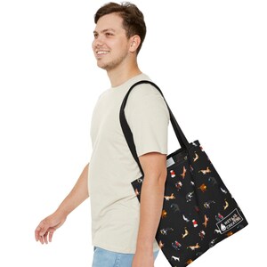 K&ouml;nnte beinhalten: Schwarze Tragetasche mit einem sich wiederholenden Muster von Hunden in verschiedenen Farben. Die Tasche hat schwarze Tr&auml;ger und den Text "WET LAB CREATIVE". Die Tasche wird &uuml;ber der Schulter getragen. Die Tasche ist aus strapazierf&auml;higem Material.