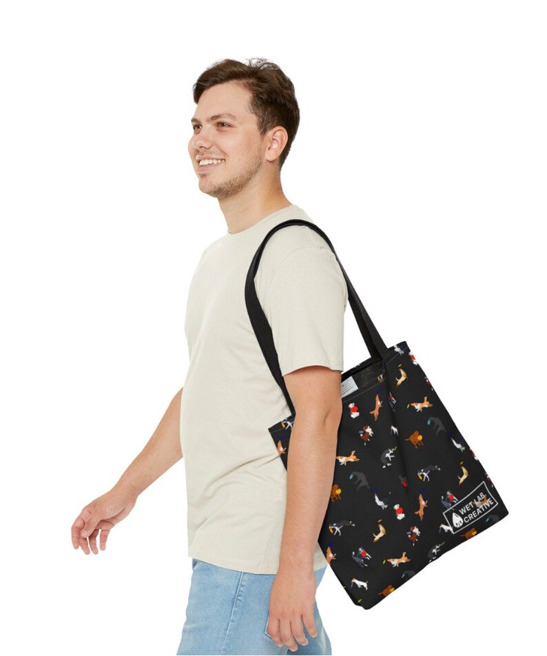 K&ouml;nnte beinhalten: Schwarze Tragetasche mit einem sich wiederholenden Muster von Hunden in verschiedenen Farben. Die Tasche hat schwarze Tr&auml;ger und den Text "WET LAB CREATIVE". Die Tasche wird &uuml;ber der Schulter getragen. Die Tasche ist aus strapazierf&auml;higem Material.