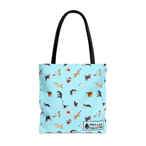 K&ouml;nnte beinhalten: Hellblaue Tragetasche mit einem sich wiederholenden Muster von spielenden Hunden. Die Tasche hat schwarze Griffe und den Text "WET LAB CREATIVE". Die Hunde sind in verschiedenen Posen, einige fangen Frisbees.